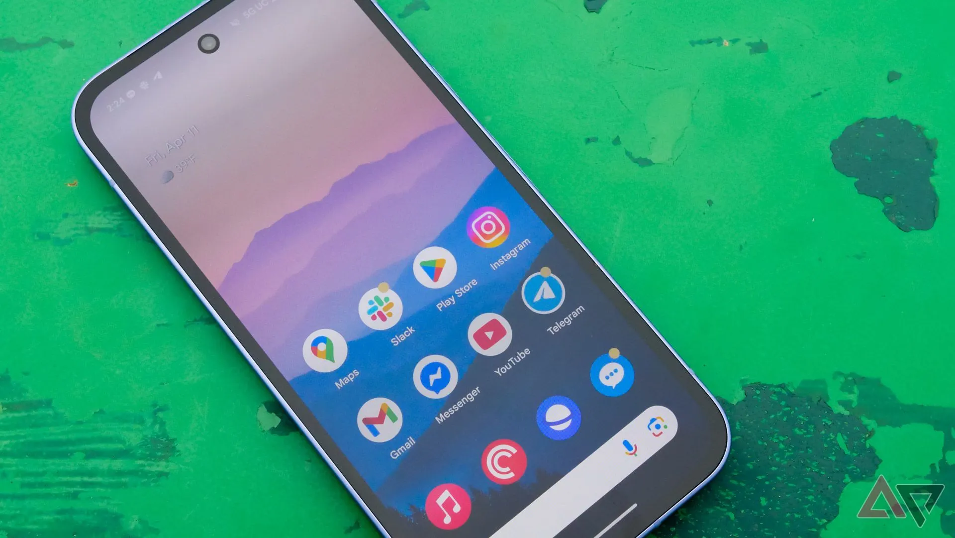 Điện thoại Google Pixel 9a đặt trên bàn xanh ngoài trời, màn hình đang bật, thể hiện khả năng hiển thị ngoài trời.