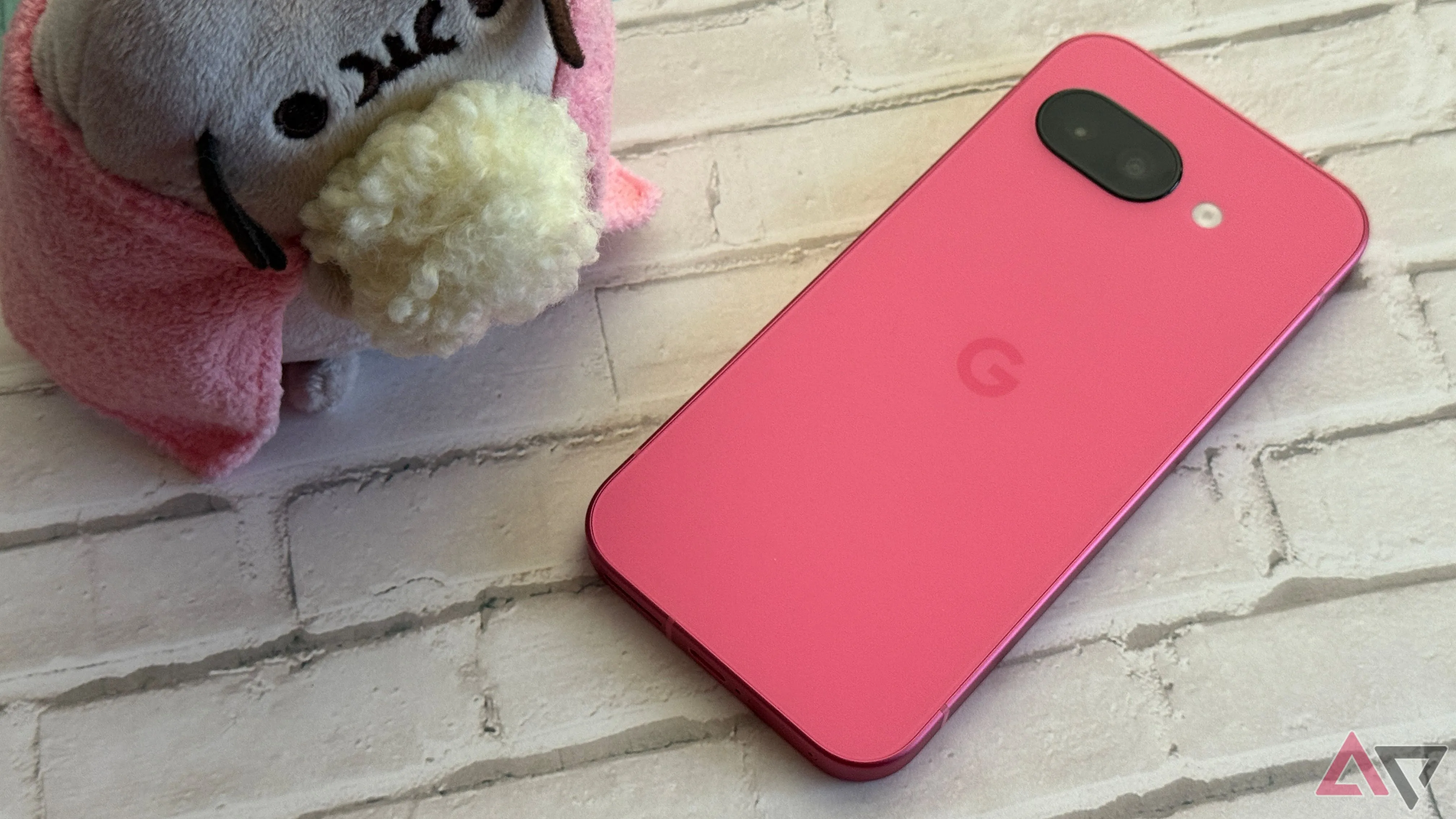 Google Pixel 9a phiên bản màu Peony đặt cạnh vật trang trí, làm nổi bật kích thước và màu sắc của điện thoại.