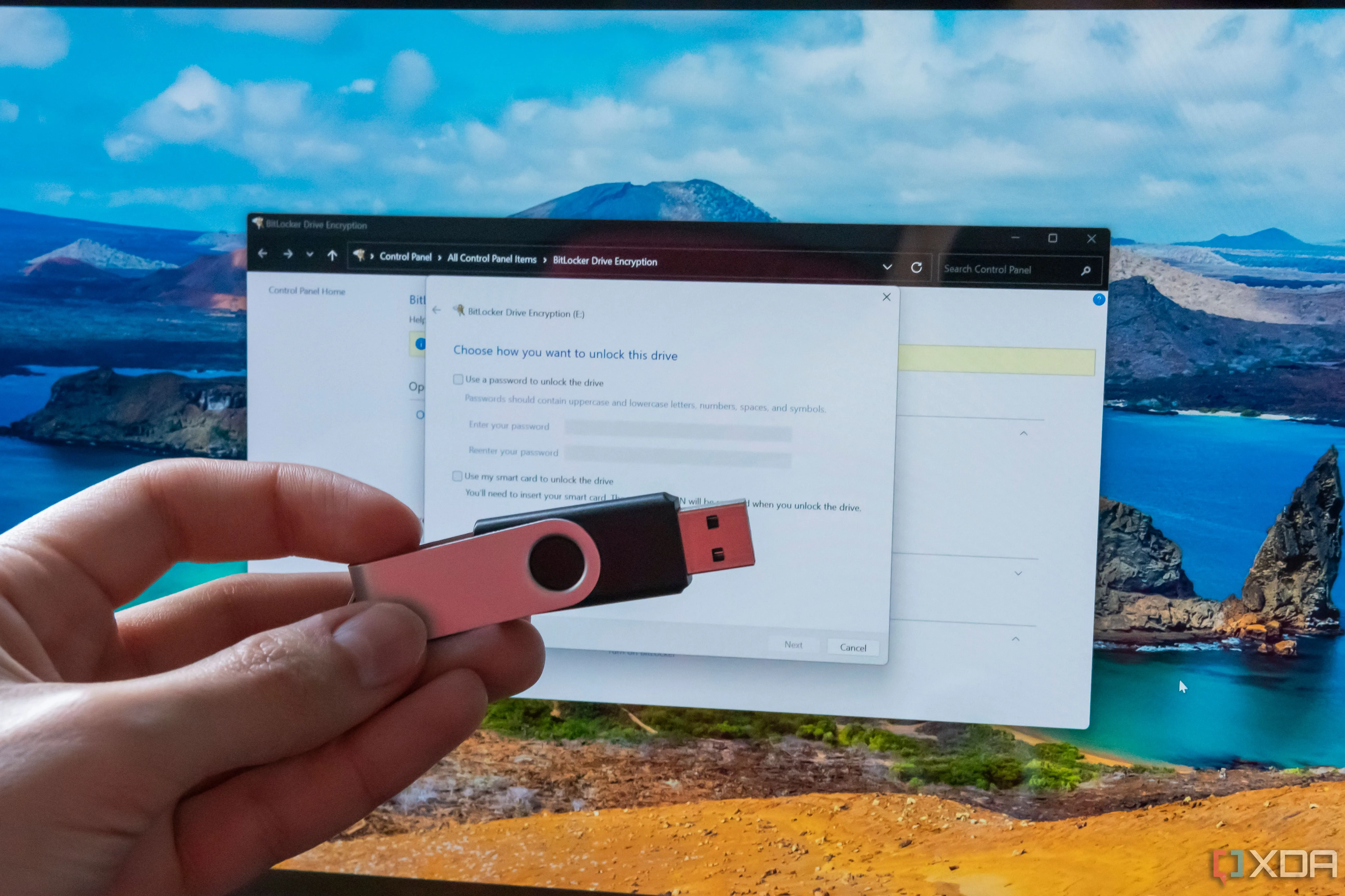 Một chiếc USB được cầm trên tay, phía sau là màn hình máy tính hiển thị cài đặt BitLocker, gợi ý các lựa chọn tạo khóa bảo mật.