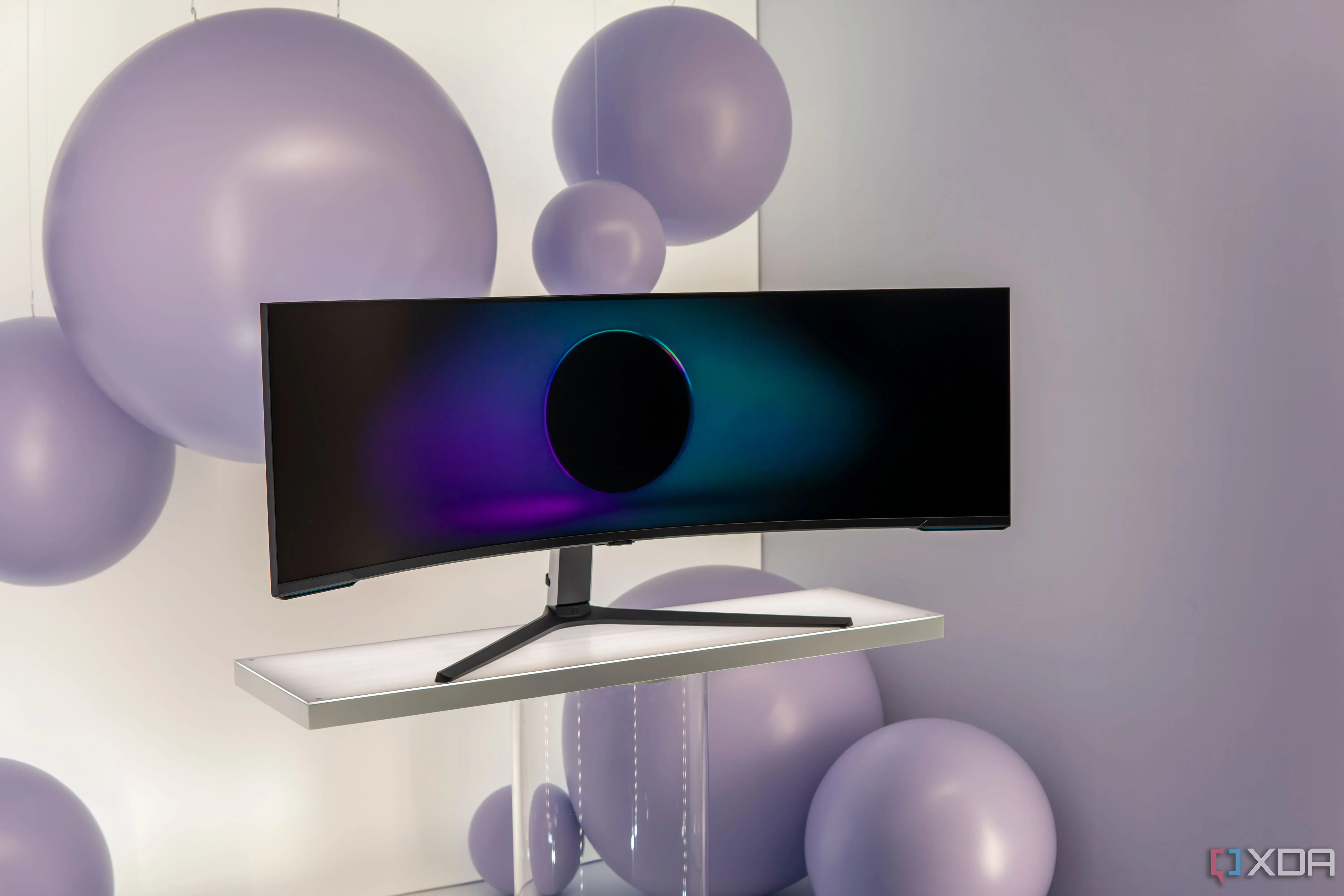 Màn hình Samsung Odyssey Neo G9 57 inch với độ cong ấn tượng, đặt trên chân đế màu trắng và bóng bay tím làm nền, thể hiện sự sang trọng của màn hình super ultrawide cao cấp.